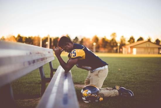 Young man in a football uniform praying at the bleachers