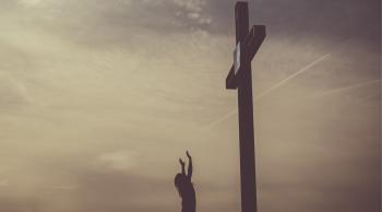 A silhouette of a woman outside with arms raised standing at the foot of a large cross