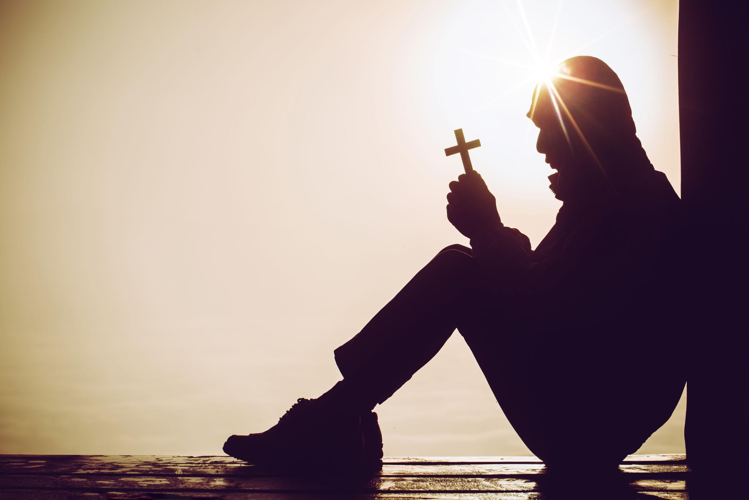 A man's silhouette in the sun, sitting against a wall and holding a cross. A glimpse of Christian persecution. 