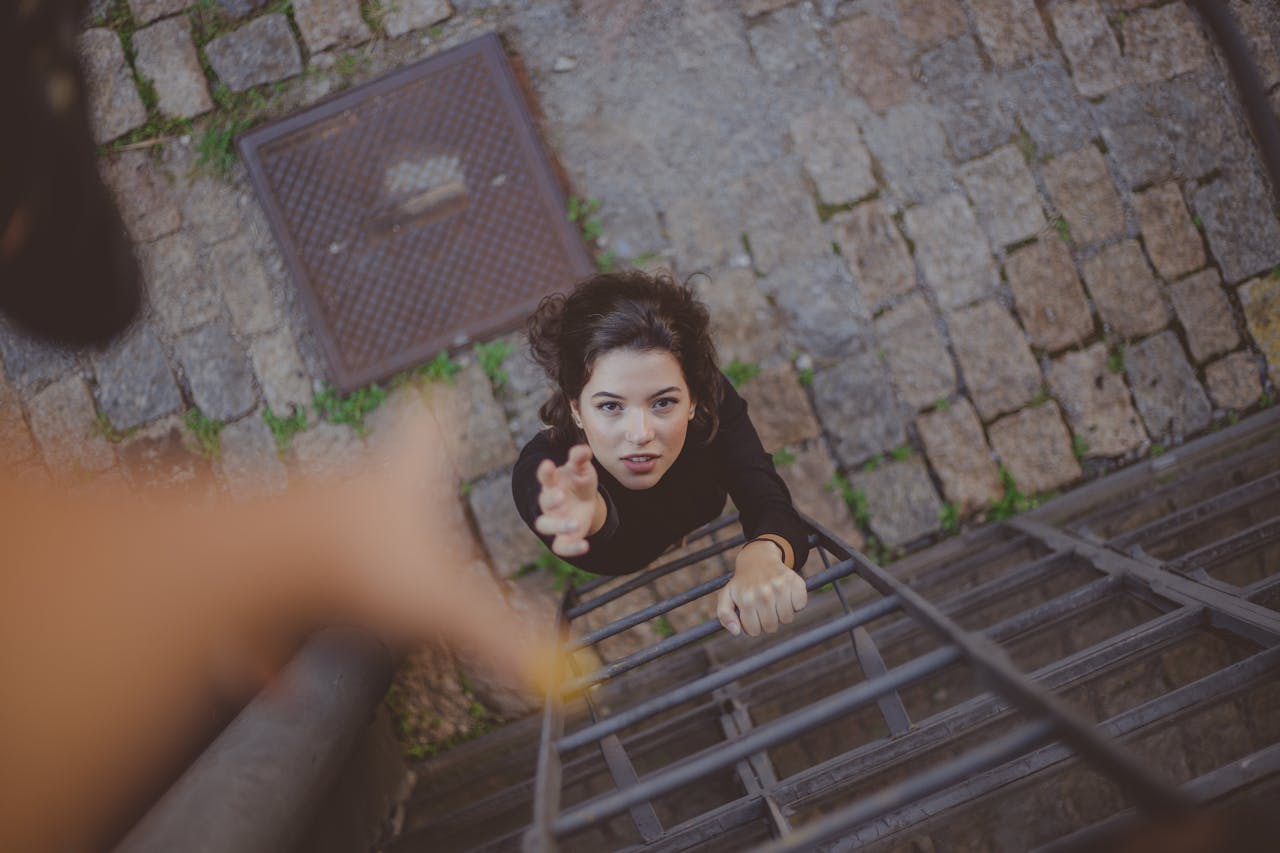 A woman at the foot of a ladder reaching up toward an open hand for help.