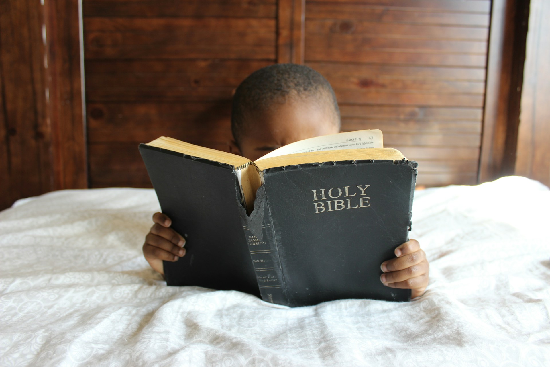 Child in bed reading a Bible held up in front of his face